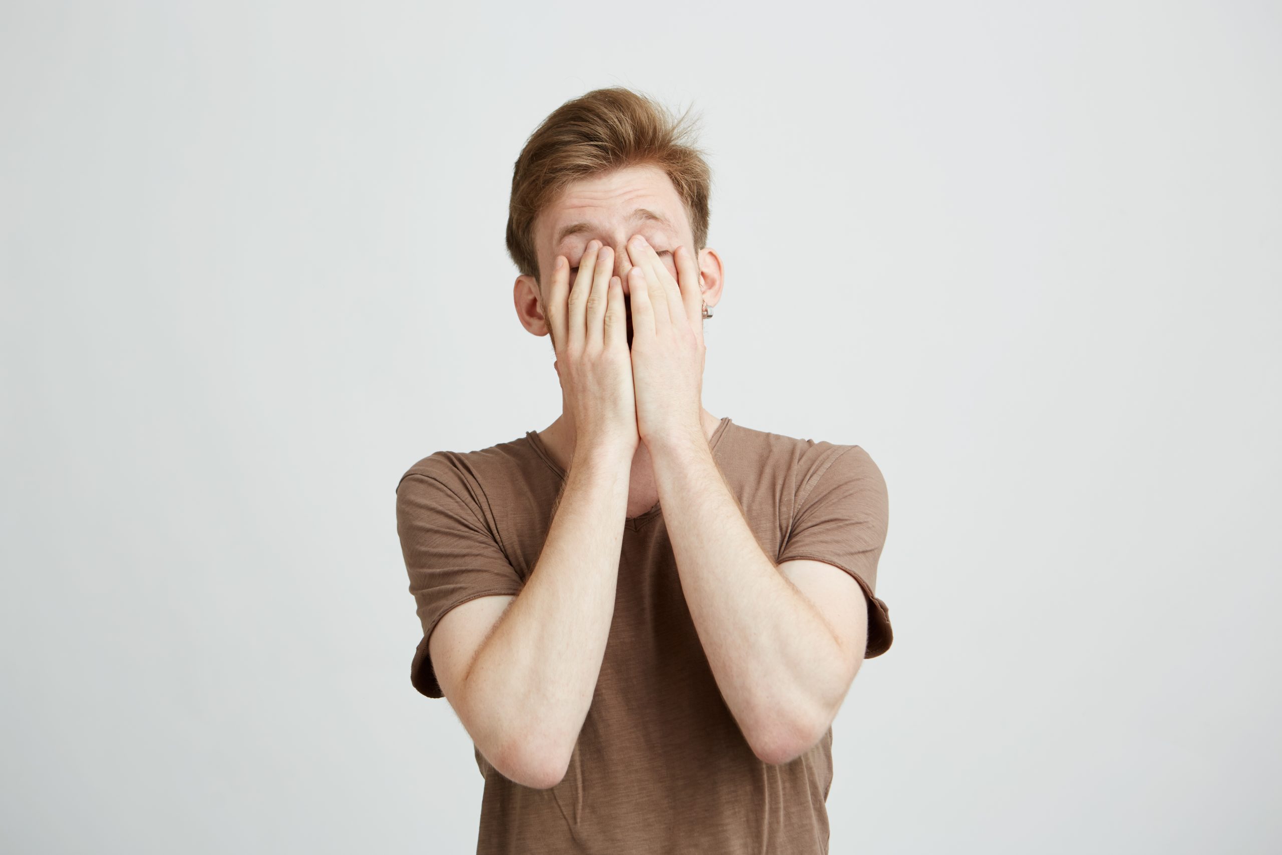 Portrait of young blond man closing face with hands over white background.